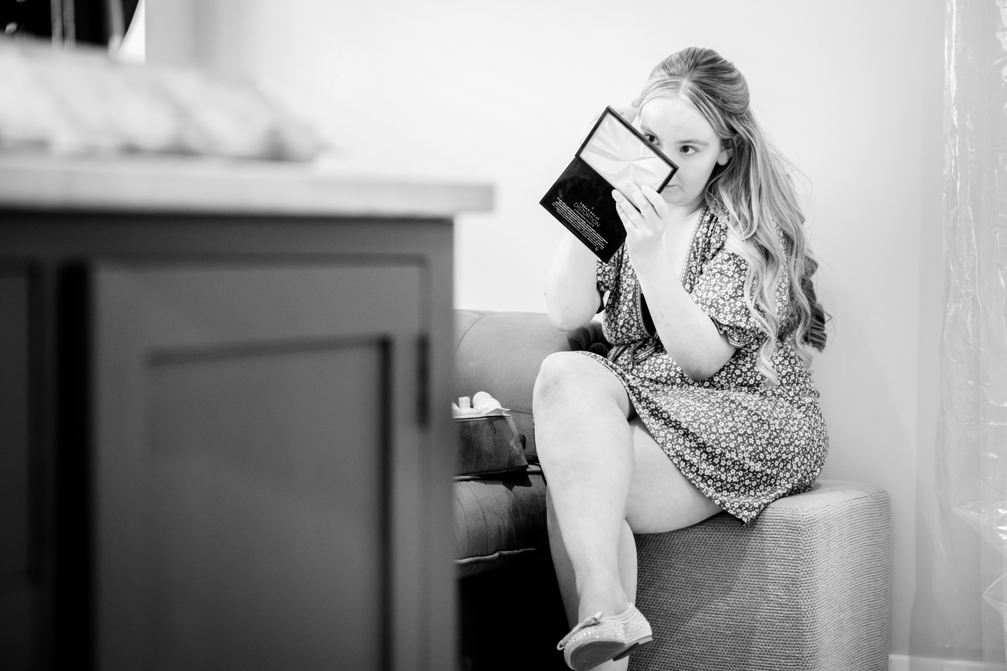 A black and white photo of a bridesmaid sitting on a couch, applying her makeup in the dressing room at Hanbury Barns.