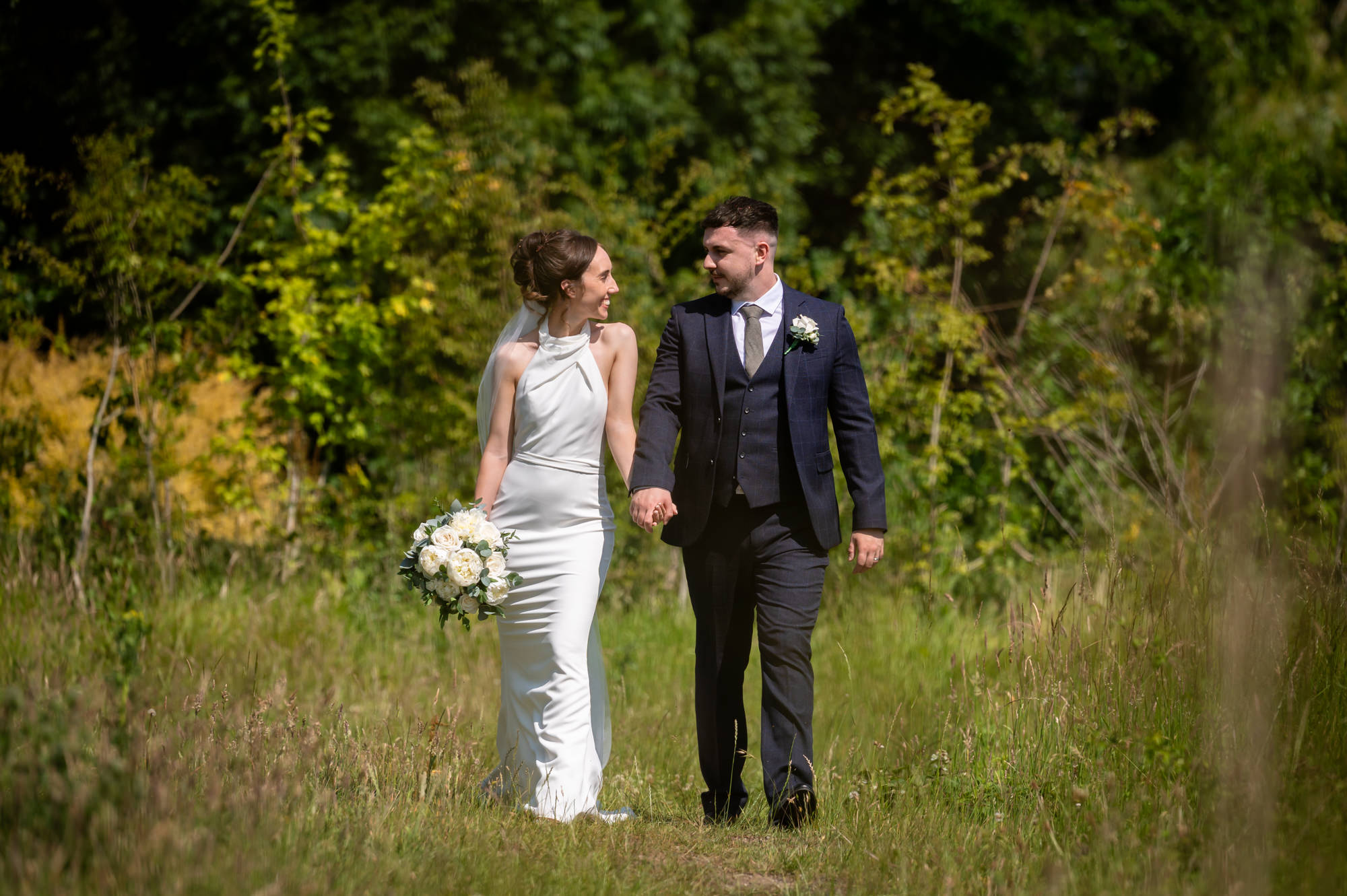A full-length portrait photo of the bride and groom walking together in a green, leafy background at Hanbury Barns.