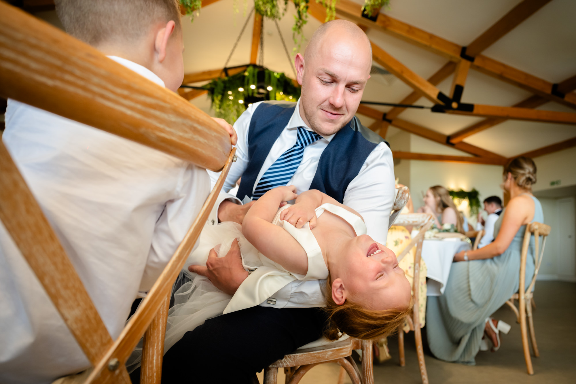 The flower girls is sat on her dad, throwing herself backwards