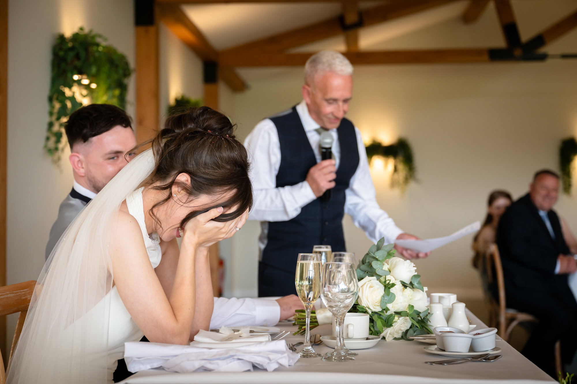 A man in a vest gives a speech, while the bride and groom are seated at the table at Hanbury Barns.