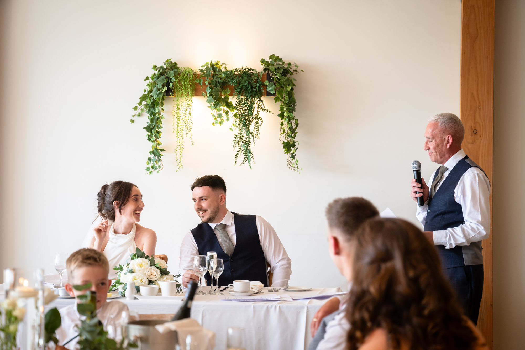 A man in a black vest and tie gives a speech to the bride, groom, and guests at Hanbury Barns.