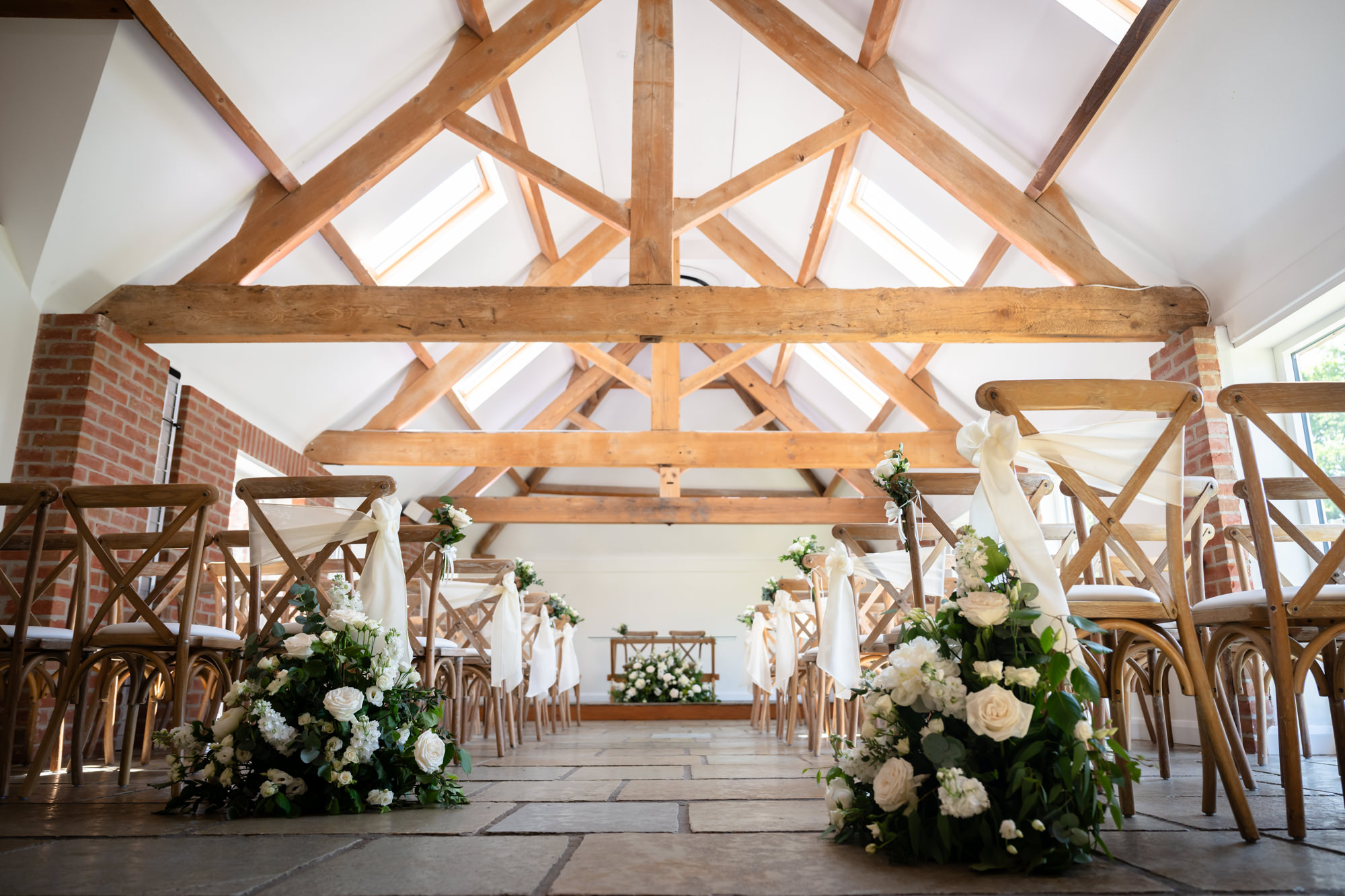 A wide-angle view of the wedding ceremony room at Hanbury Barns, decorated with white flowers and rustic chairs.