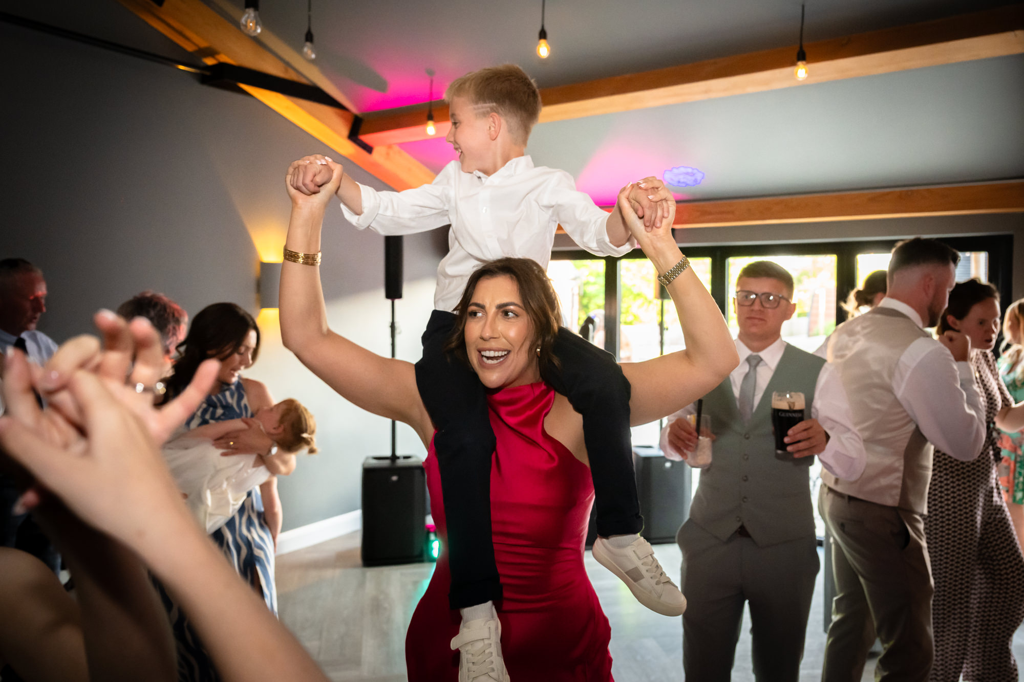 A woman is laughing with a boy on her shoulders during the wedding party at Hanbury Barns.
