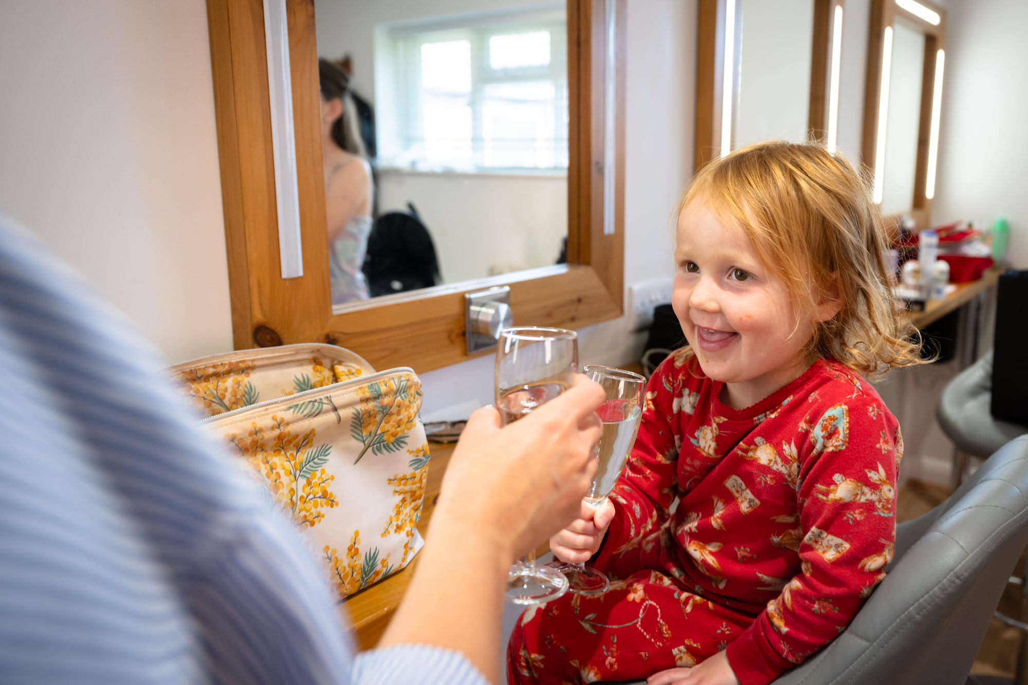 A young girl in a red and white pyjama smiles as she holds up two glasses of champagne with an adult in the dressing room at Hanbury Barns.