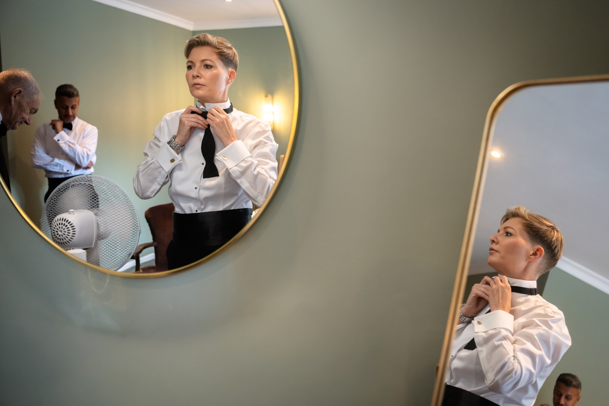 Multiple reflections in mirrors show the bride in the white tuxedo jacket adjusting her bow tie at Springbank Manor.