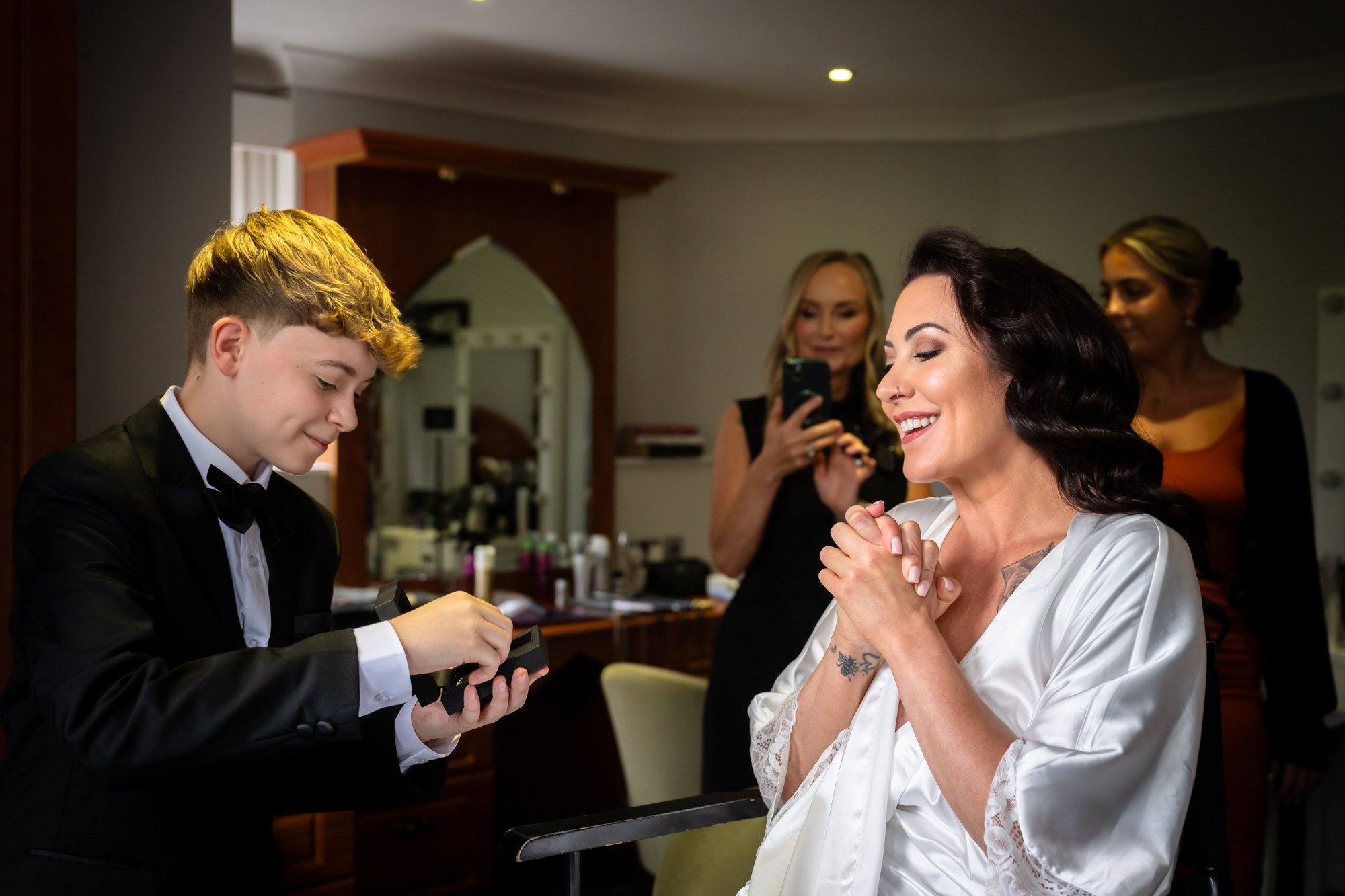 The bride in a white dressing gown laughs as her son, in a tuxedo, gives her a gift at Springbank Manor.