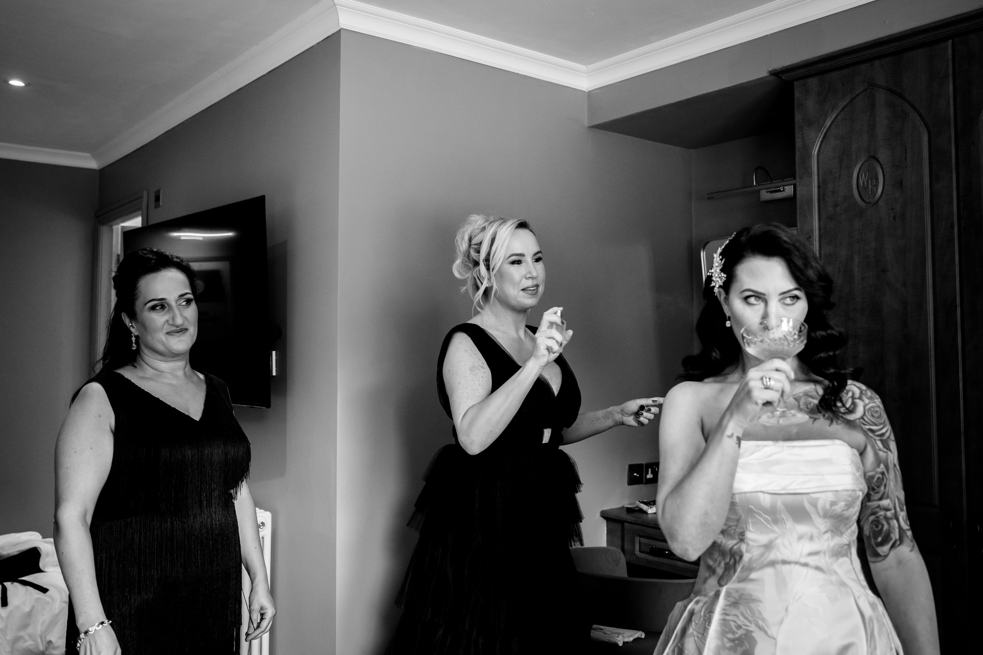 A black and white photo of a bridesmaid spritzing perfume on the bride while she is sipping champagne at Springbank Manor.