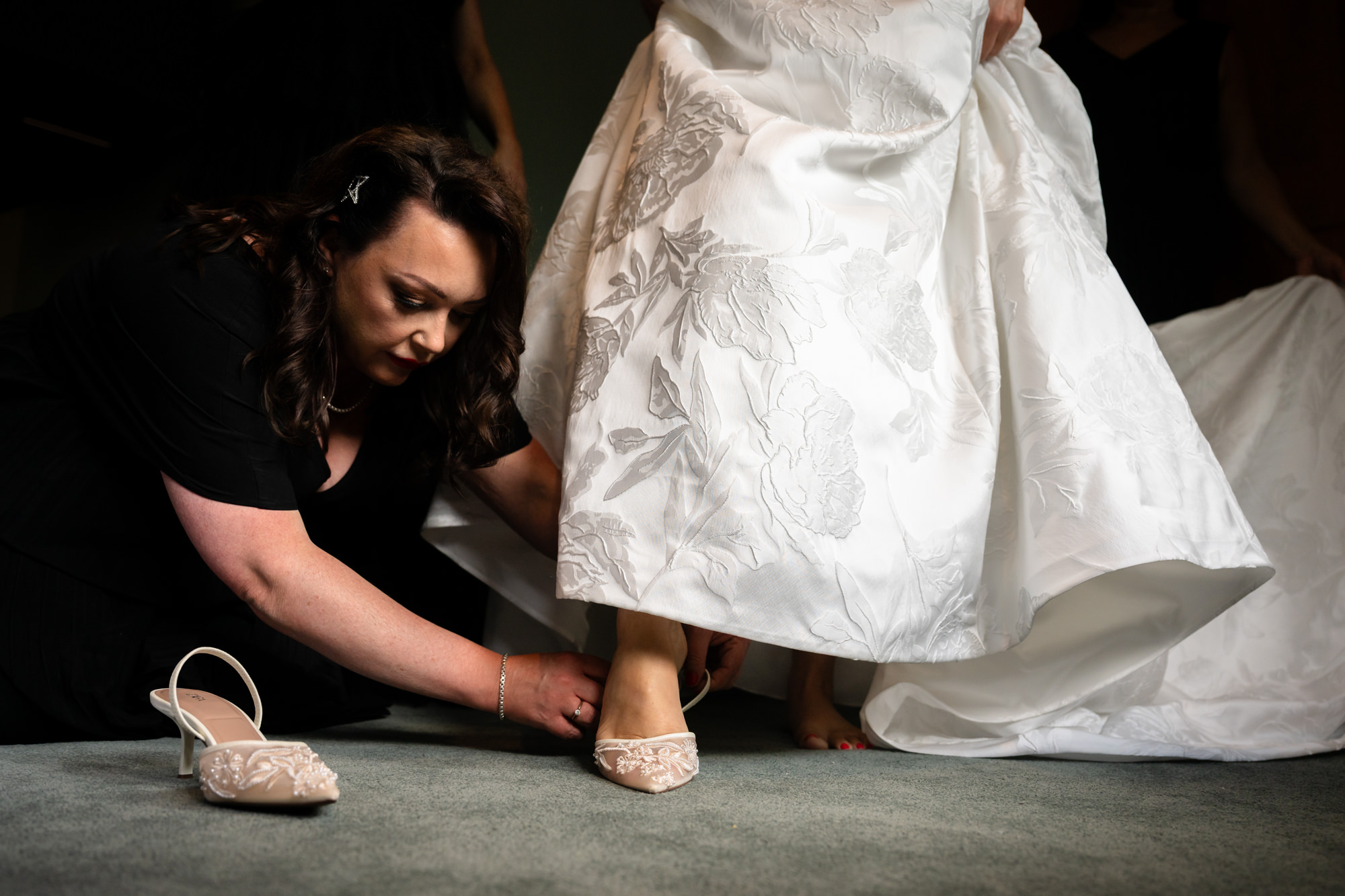 A woman in a black dress helping the bride in the white dress with her shoes at Springbank Manor.