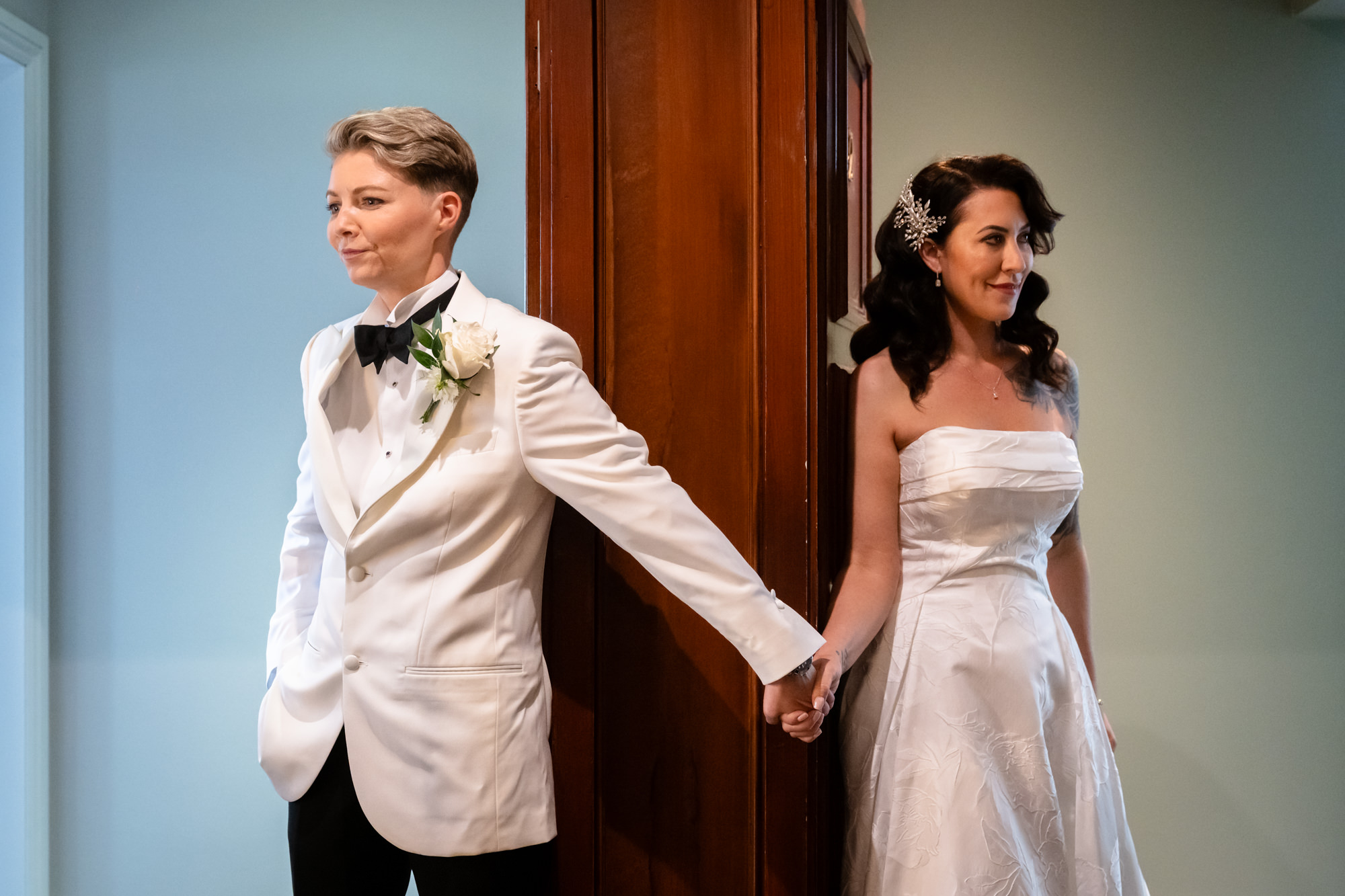 A bride in a white tuxedo and another bride in a white dress stand back to back, holding hands before the ceremony at Springbank Manor.