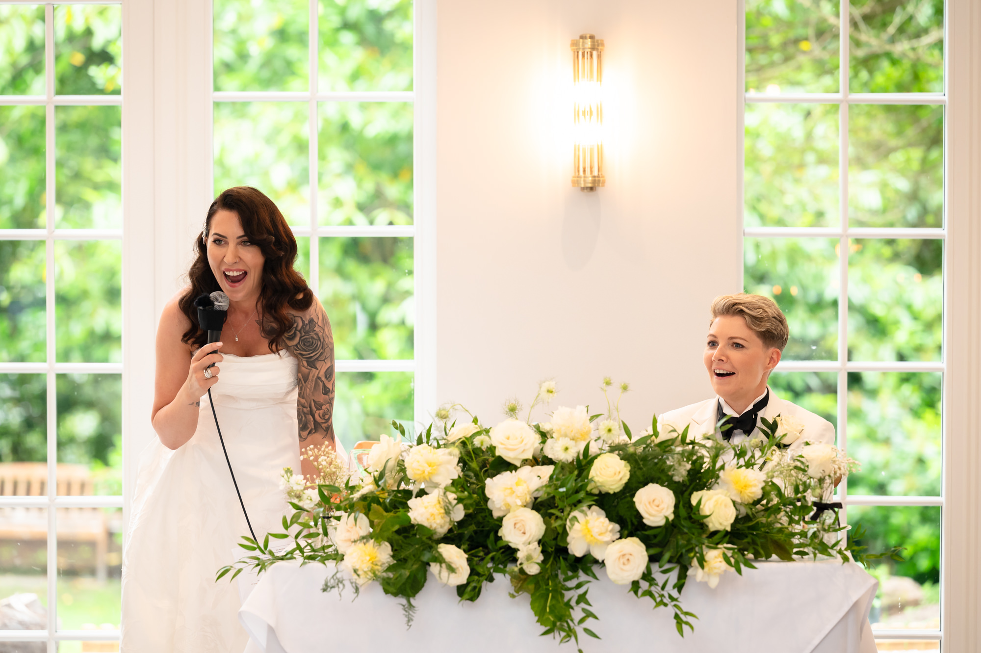 A photo of the two brides at their table, with the bride in the dress holding a microphone and the bride in the tuxedo smiling at Springbank Manor.