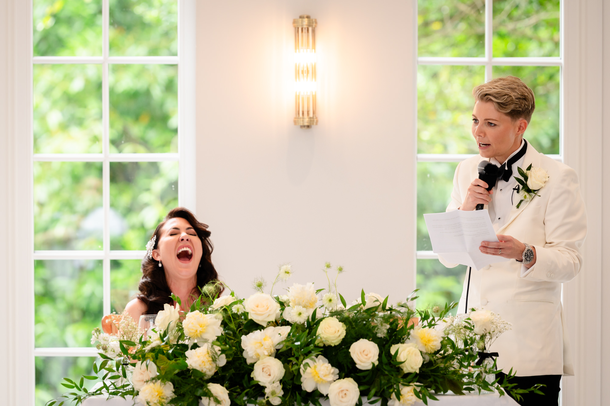 The two brides are seen in a photo at their table, with the bride in the dress laughing out loud and the bride in the tuxedo holding a microphone at Springbank Manor.