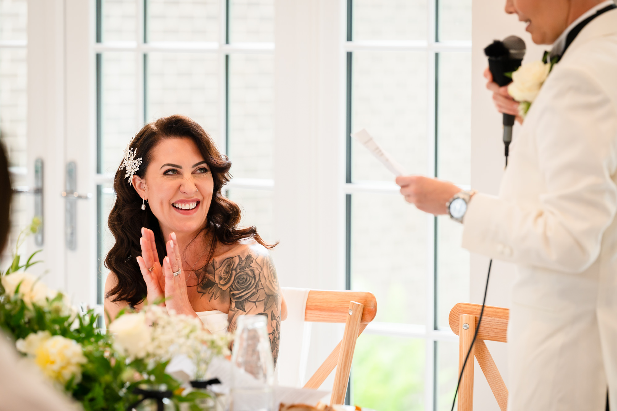 The bride in the white dress laughs as she looks at the bride in the tuxedo at their table at Springbank Manor.