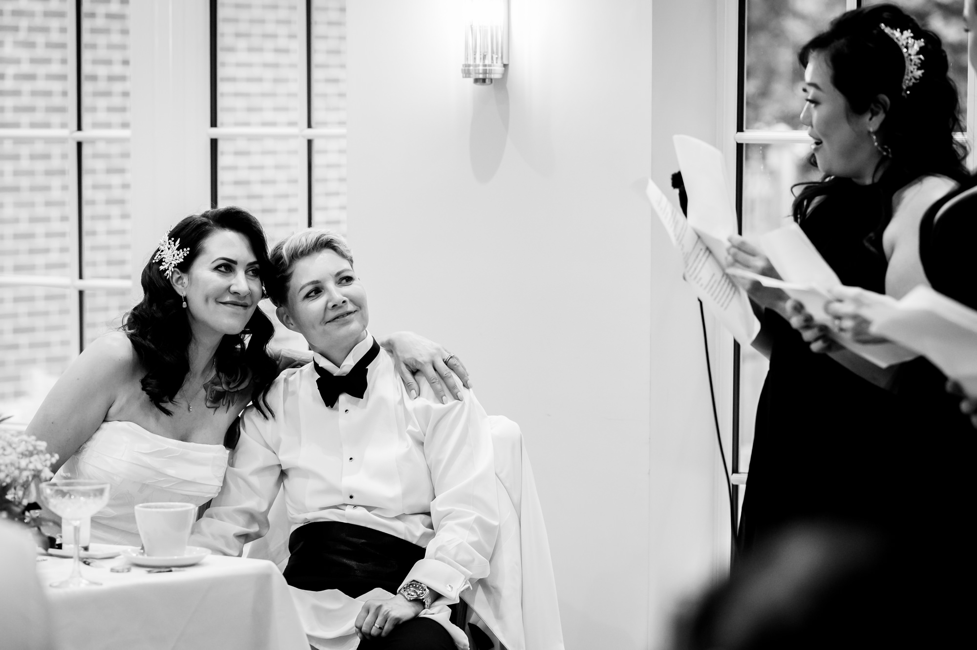 A black and white photo of the two brides at their table, looking at a woman giving a speech at Springbank Manor.