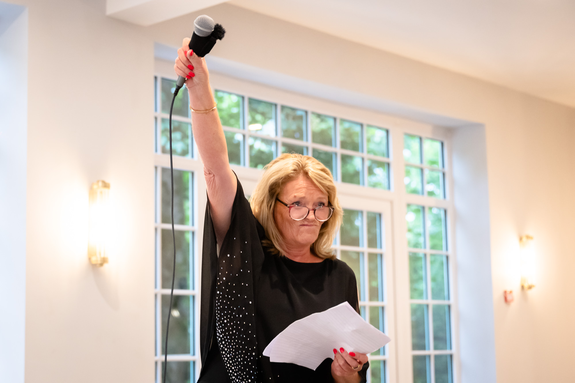 A woman in a black shirt stands with a microphone held up high in her hand at the wedding at Springbank Manor.
