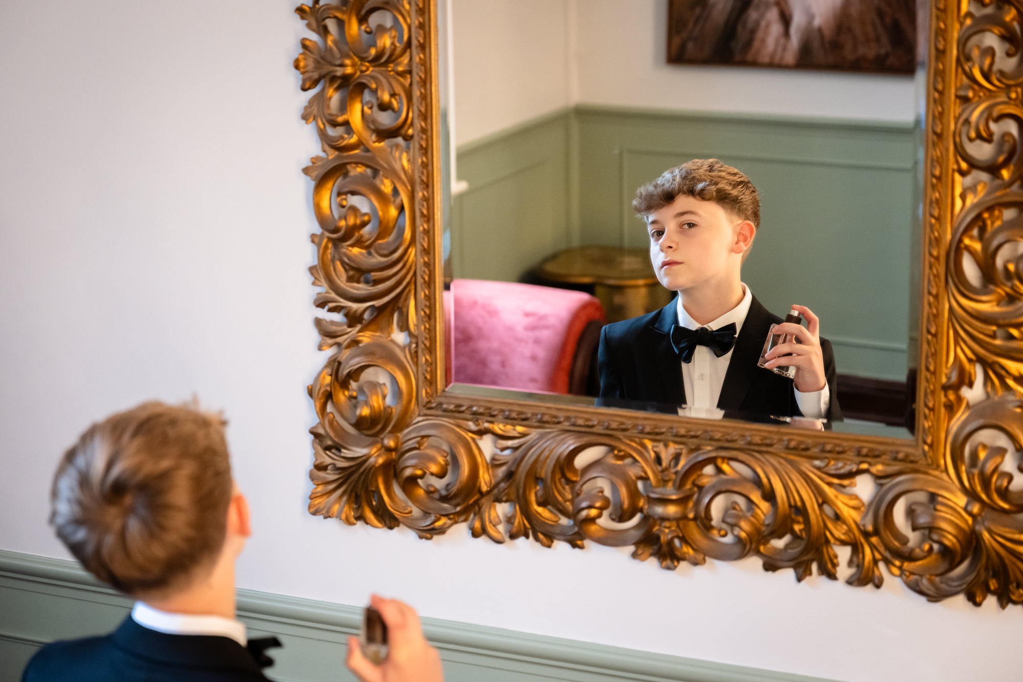 A young boy in a black tuxedo jacket with a bow tie looks at his reflection in a large, ornate golden mirror at Springbank Manor.