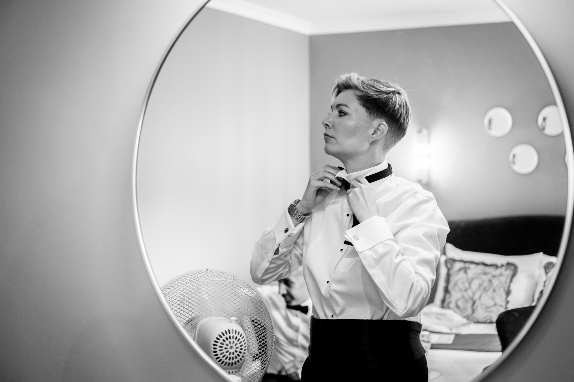 9 A black and white photo of the bride in the white tuxedo jacket looking at her reflection in a round mirror while adjusting her black bow tie at Springbank Manor.