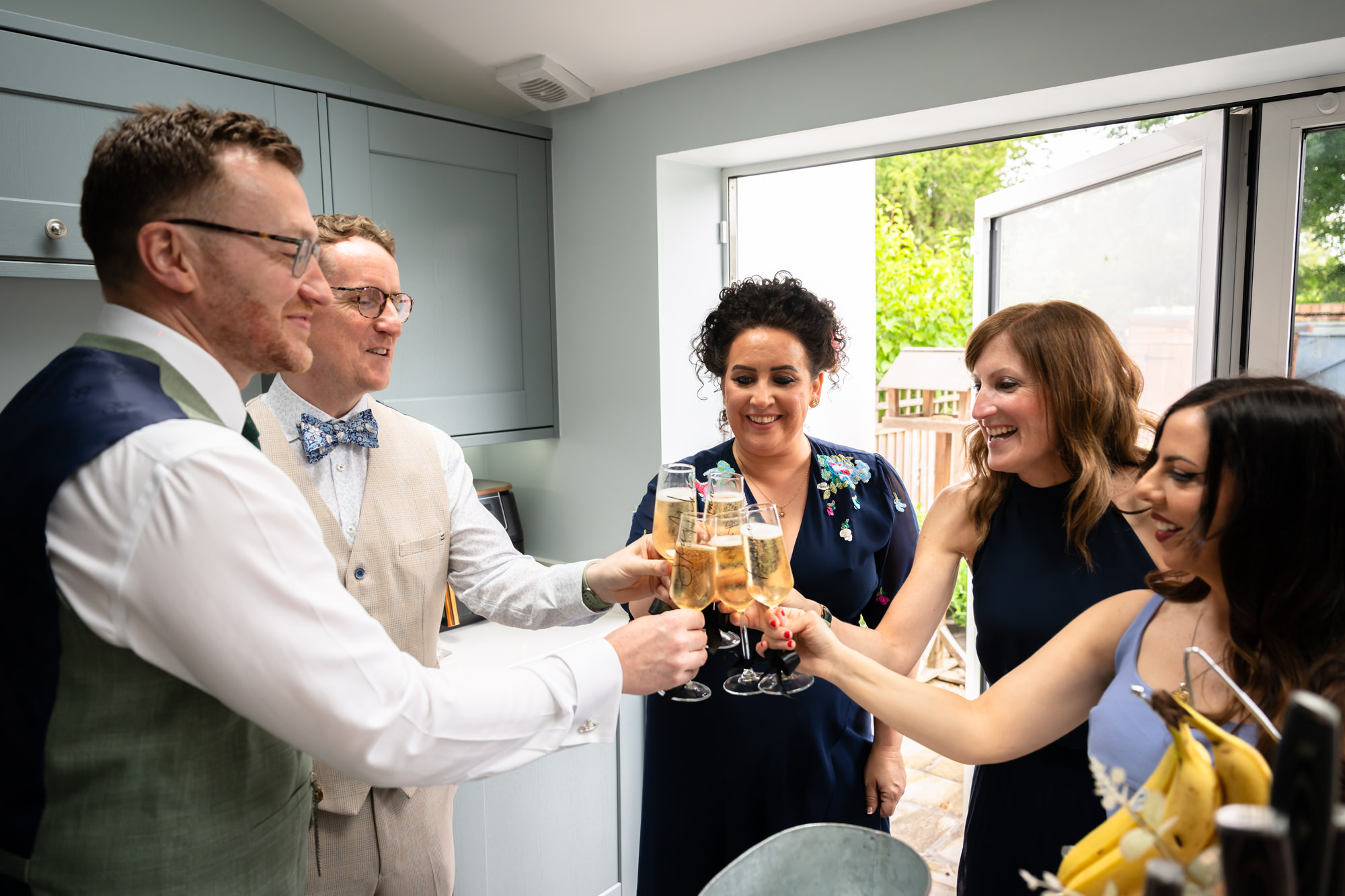 Two grooms, one wearing a beige suit with a blue bowtie and the other wearing a green suit with a green tie, toast with champagne flutes with three women in a kitchen at the grooms' house.