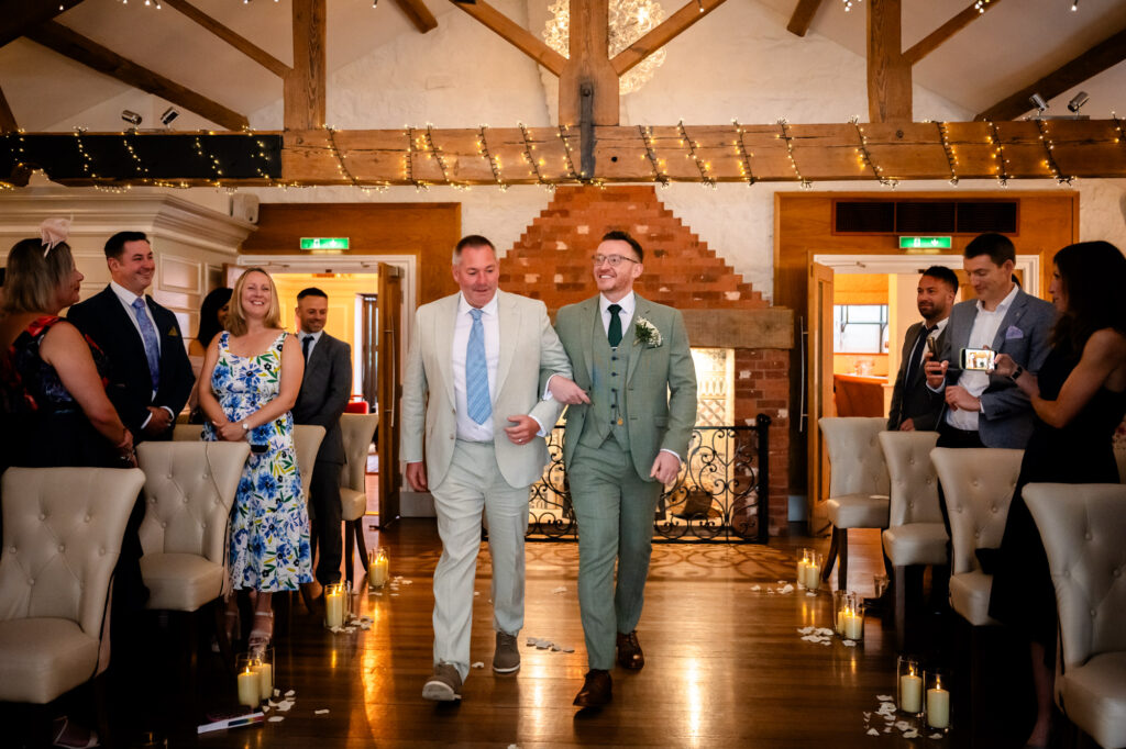 Wedding at Allerton Manor. The groom in a green suit walks down the aisle, arm-in-arm with a man in a light beige suit, as guests watch from the chairs lining the aisle.