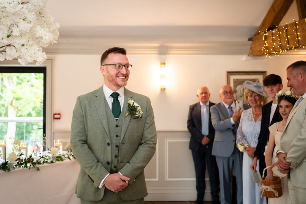 The groom in a green suit stands smiling at the end of the aisle during the ceremony.