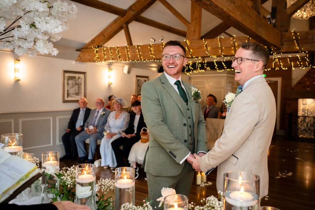 Both grooms stand at the front of the room during the ceremony, smiling at each other.