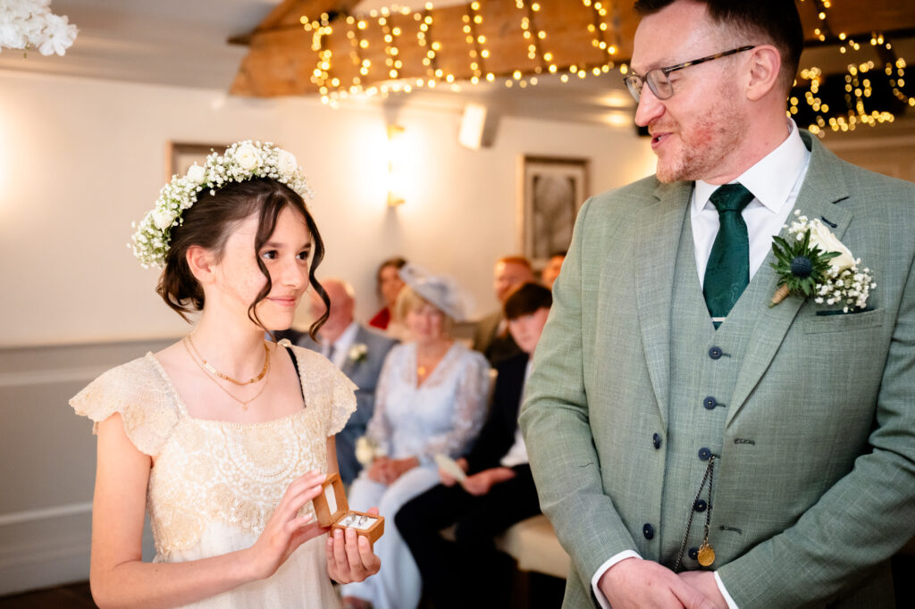 The groom in the green suit stands smiling next to a young girl in a pink dress and a floral headband.