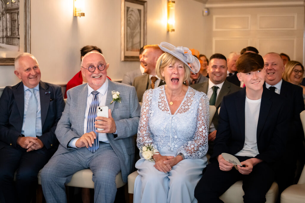 Guests sit in the front row, watching the ceremony, including a woman with a light blue hat and a man with a grey suit.