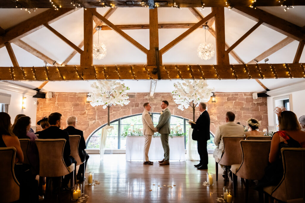 A wide shot of the grooms and the officiant at the front of the room, with guests seated on both sides of the aisle.