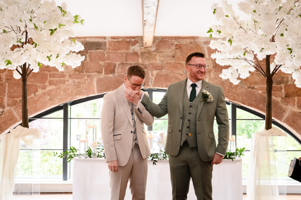 The grooms are at the front of the room, one in a beige suit and the other in a green suit, with one holding the hand of the other.