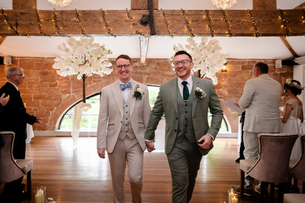 The grooms walk hand-in-hand down the aisle together after their ceremony, smiling at the guests.