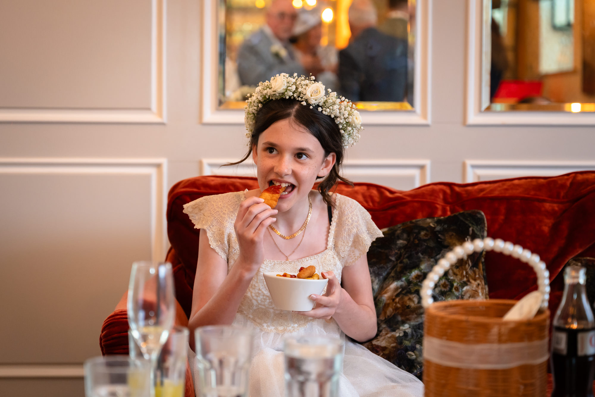 A young girl in a pink dress and floral headband sits smiling while eating something.