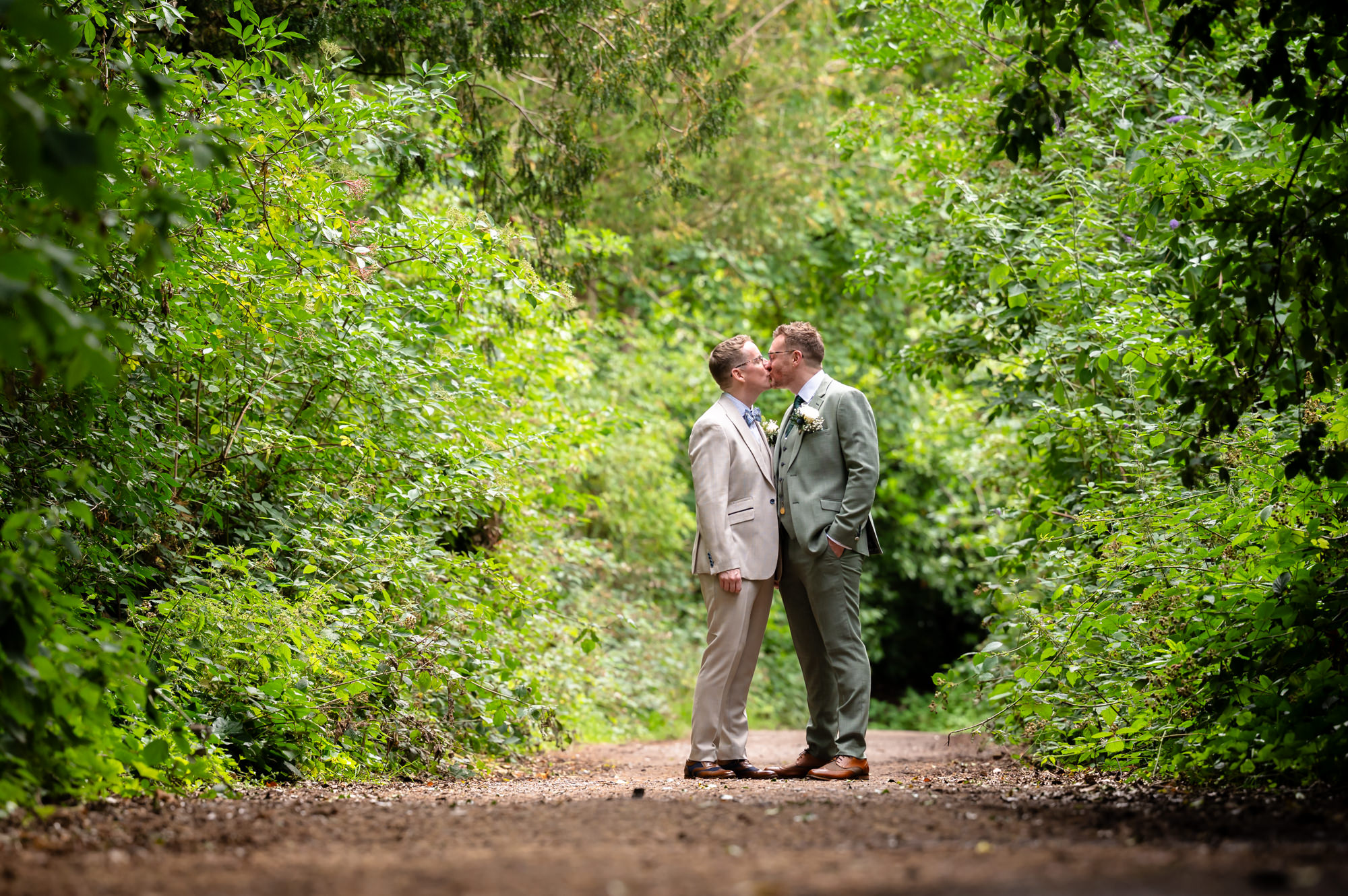 A portrait photo of the grooms, one in a green suit and the other in a beige suit, standing outdoors with green foliage in the background.