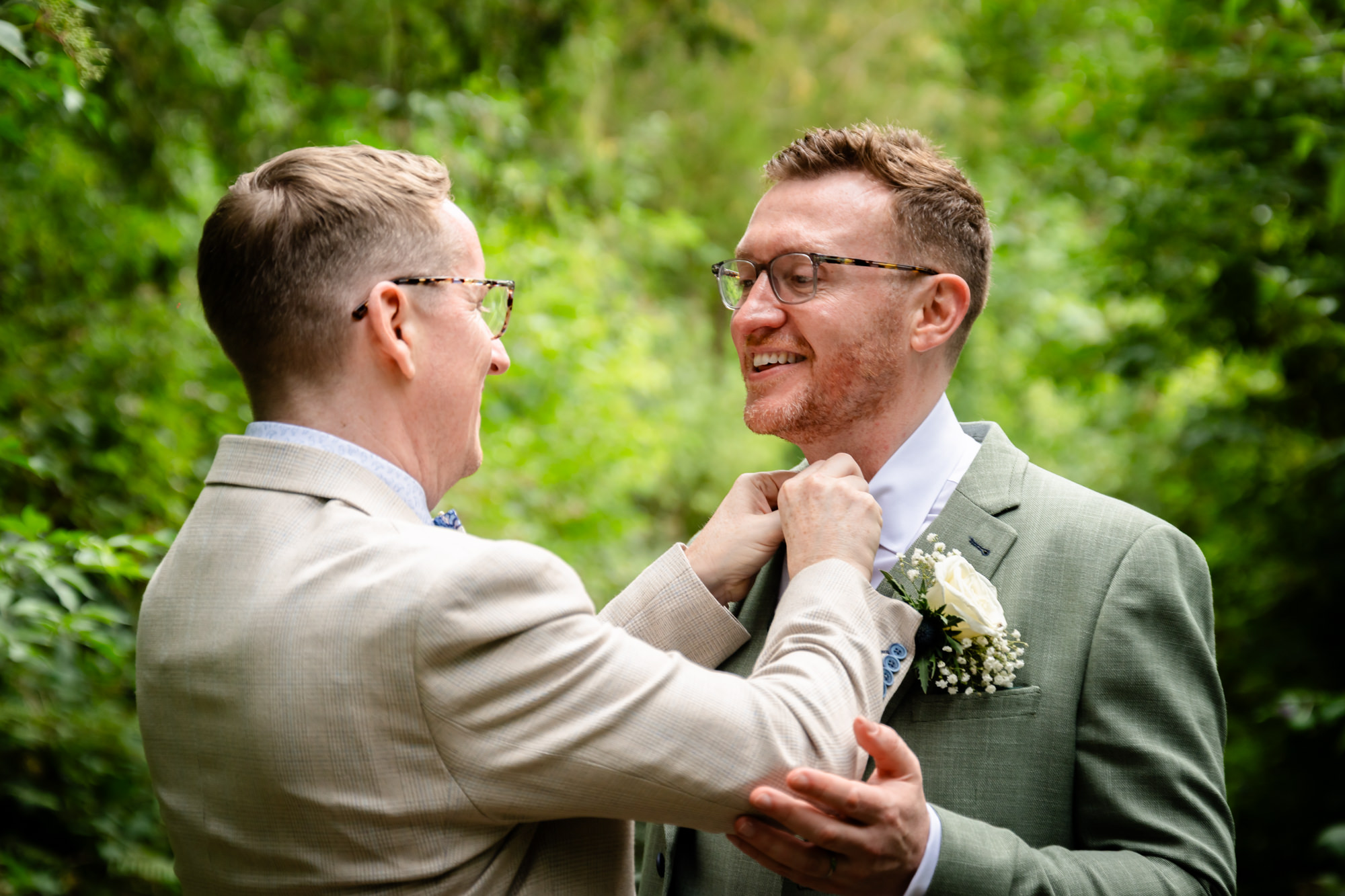 A portrait of the grooms, one in a beige suit and the other in a green suit, adjusting his tie