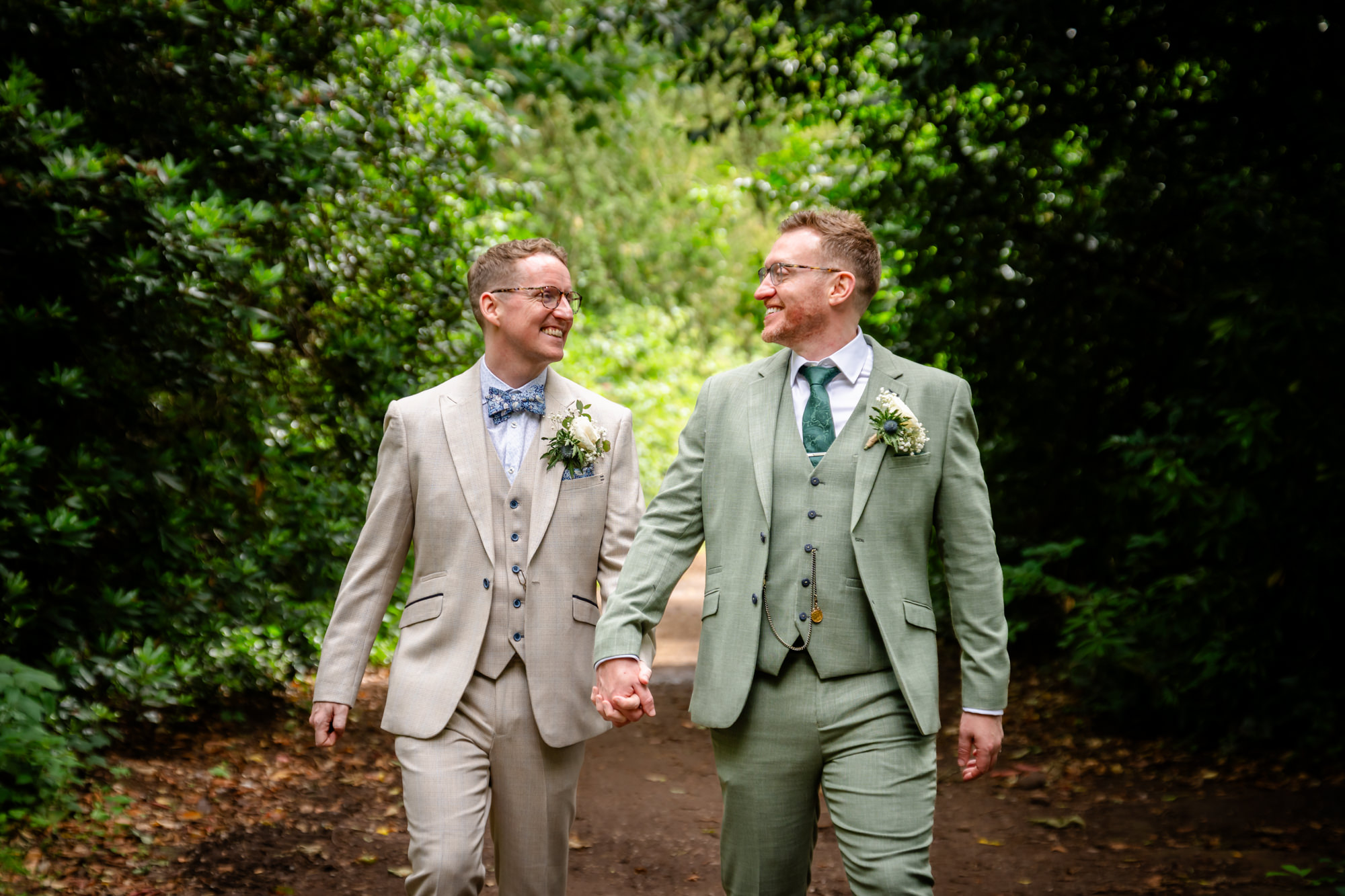 A portrait of the grooms, one in a beige suit and the other in a green suit, walking hand-in-hand down a shaded path with trees on either side.