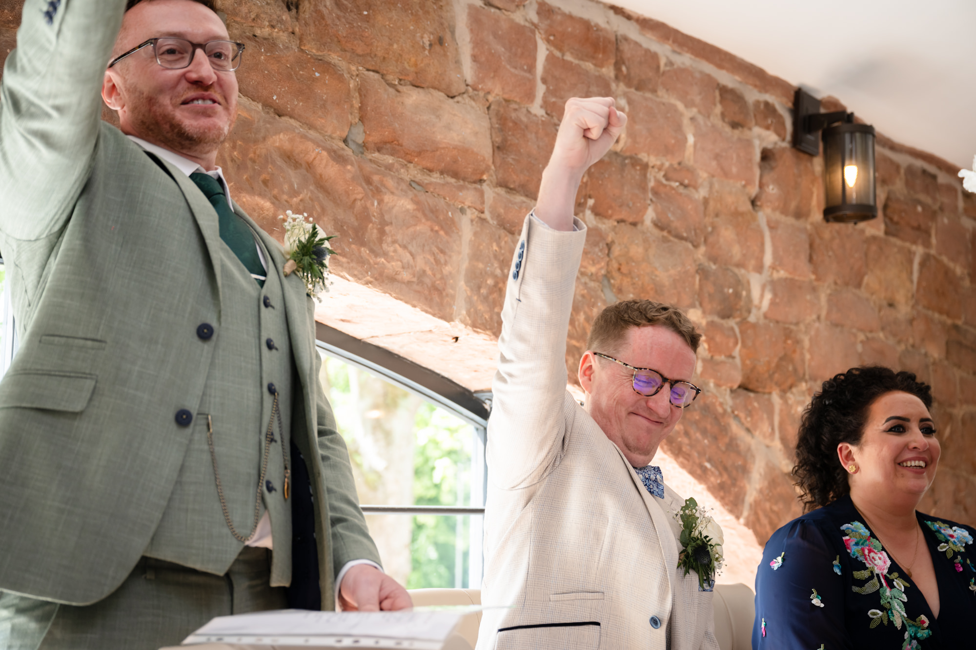The groom in a beige suit sits and listens to the speeches with a woman next to him who is also listening.