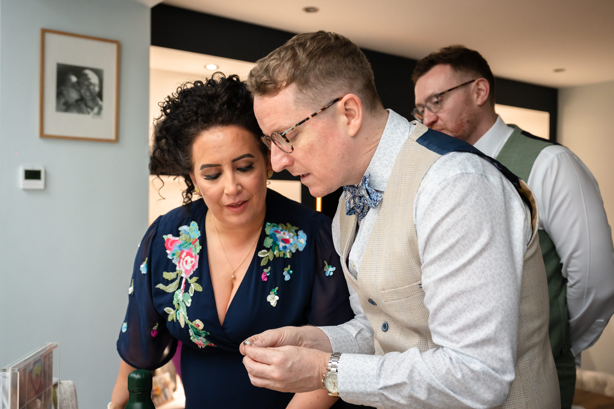 A groom in a beige suit with a blue bowtie shows a wedding ring to a woman with curly hair in a navy dress, as the other groom in a green suit looks on from behind at the grooms' house.
