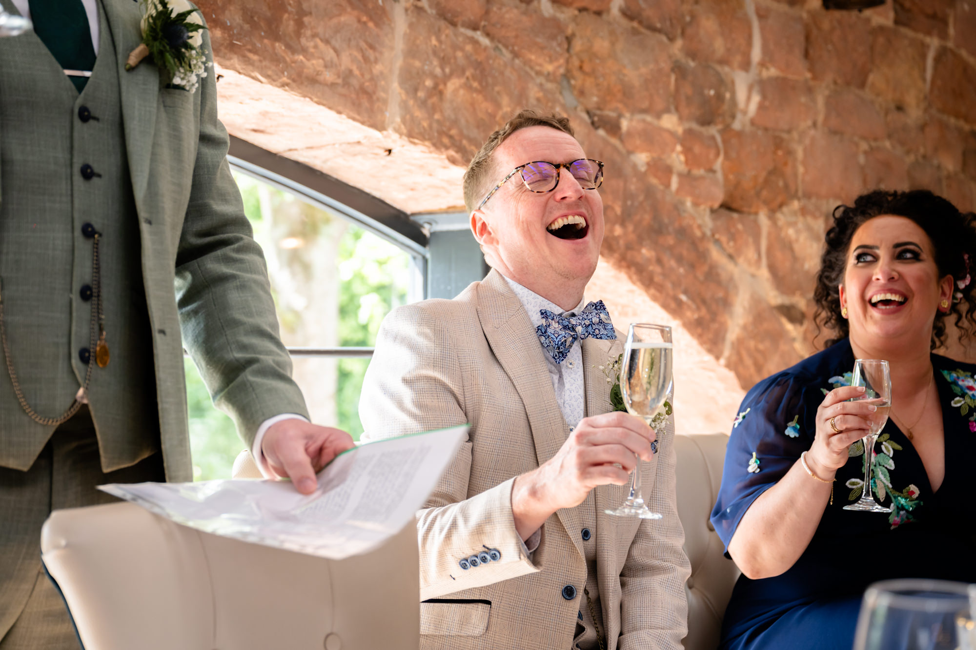 The groom in a beige suit laughs heartily while holding a glass of wine, during his husband's speech