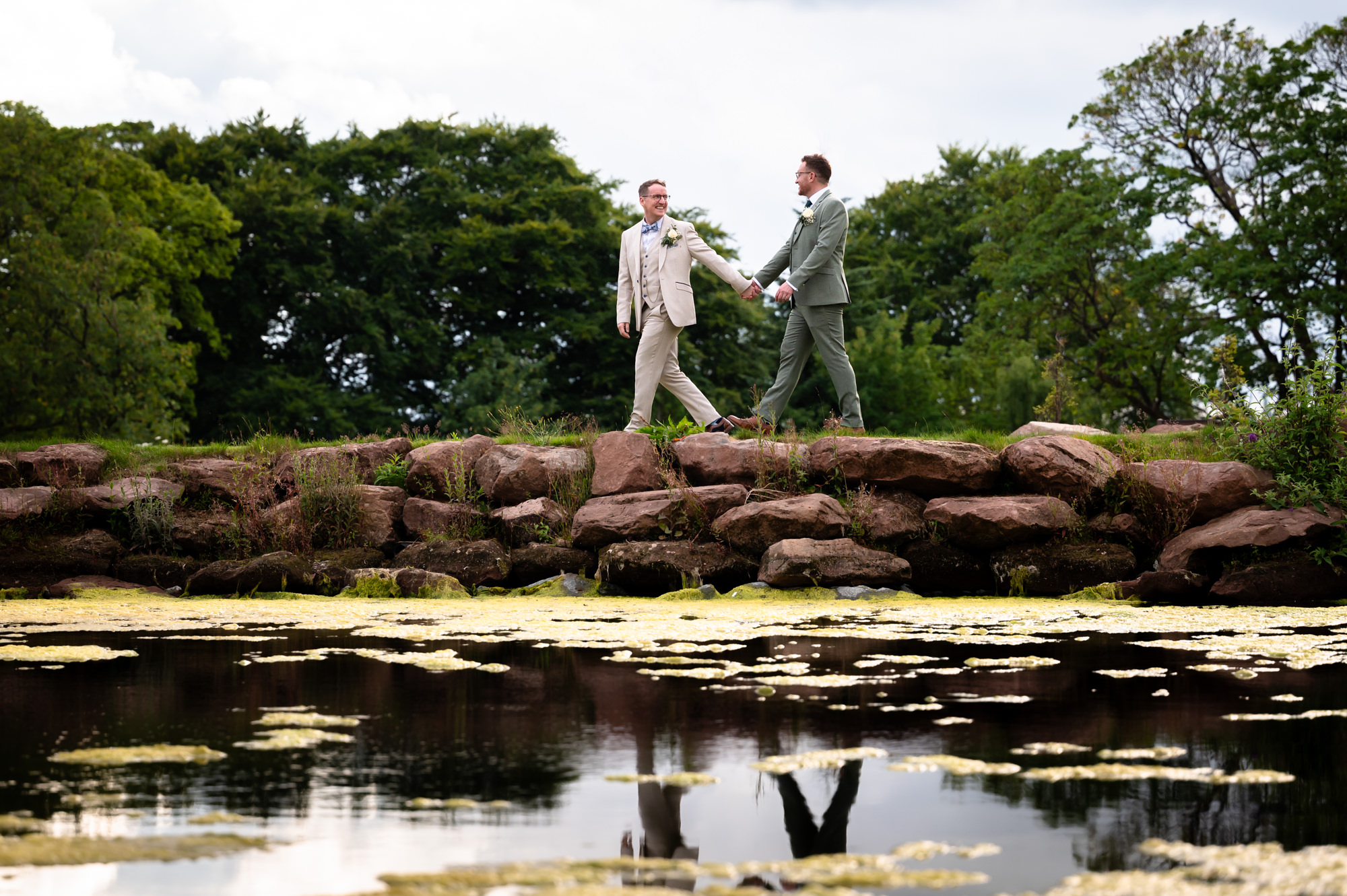 A wide-angle portrait photo of the grooms walking together on the grass near a pond, with a tree line in the background at Allerton Manor.