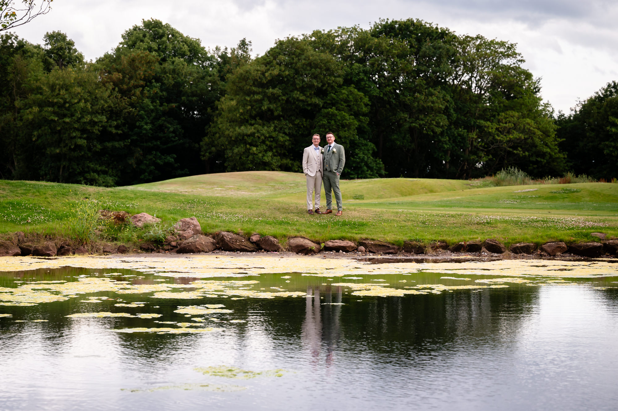 A wide-angle portrait photo of the grooms walking together on a path with a large pond with lily pads in the foreground.
