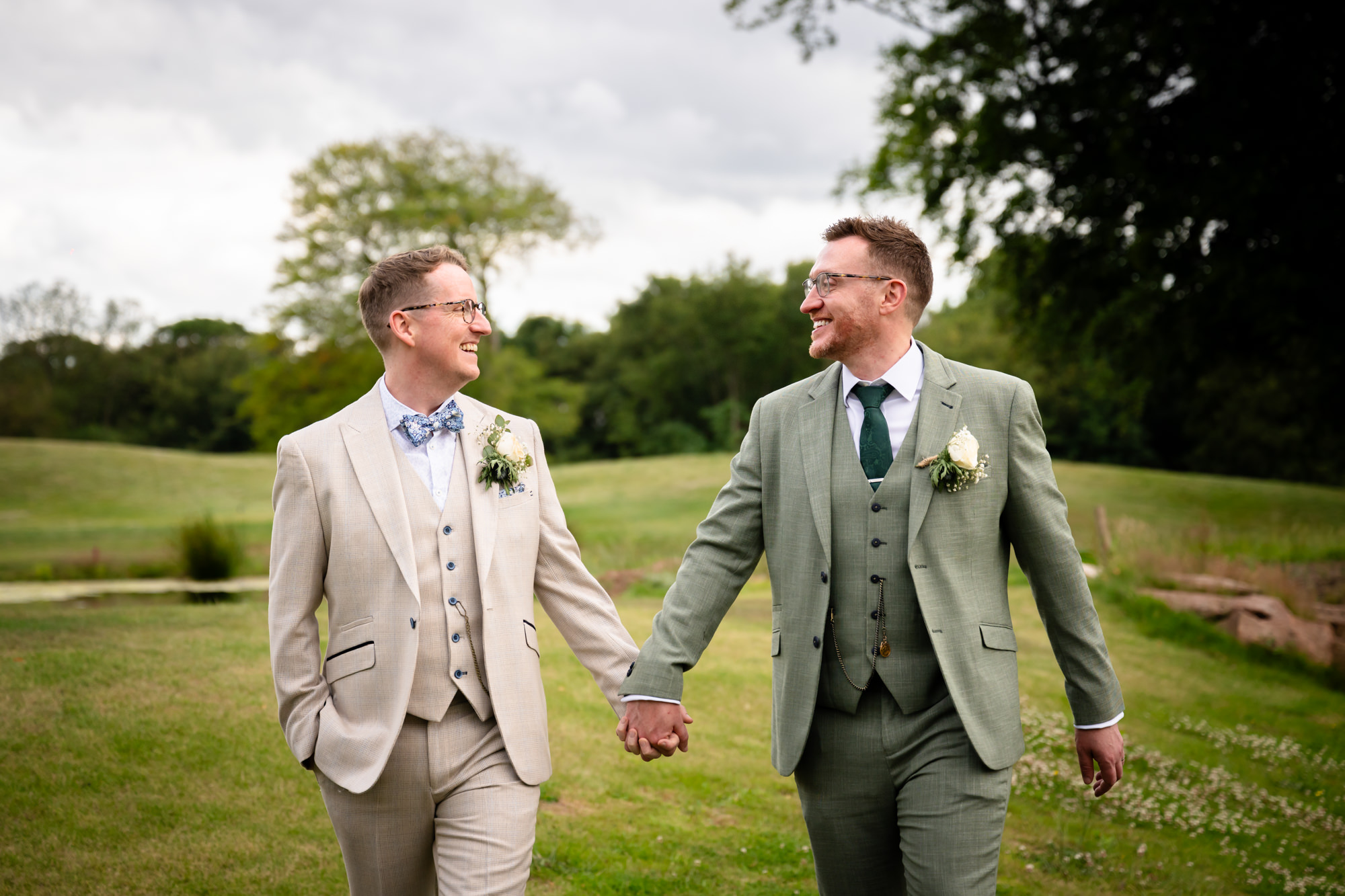 A portrait photo of the grooms holding hands and smiling at each other while walking on a grassy lawn with trees in the background.
