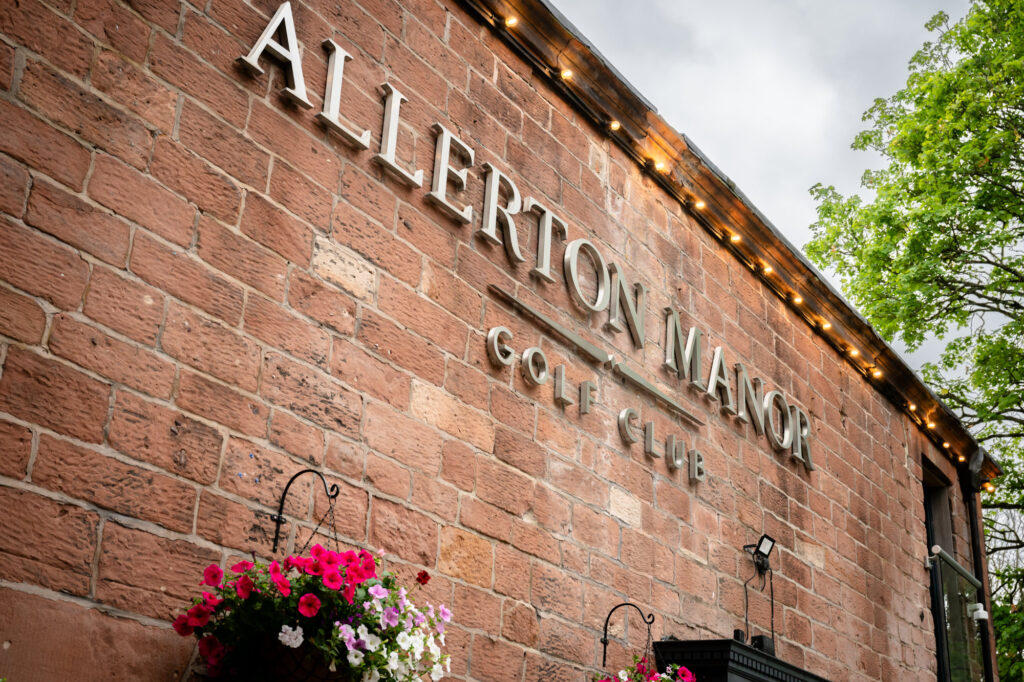 A close-up shot of the brick exterior of the Allerton Manor Golf Club, featuring the venue's large metal sign with hanging baskets of pink and white flowers below.