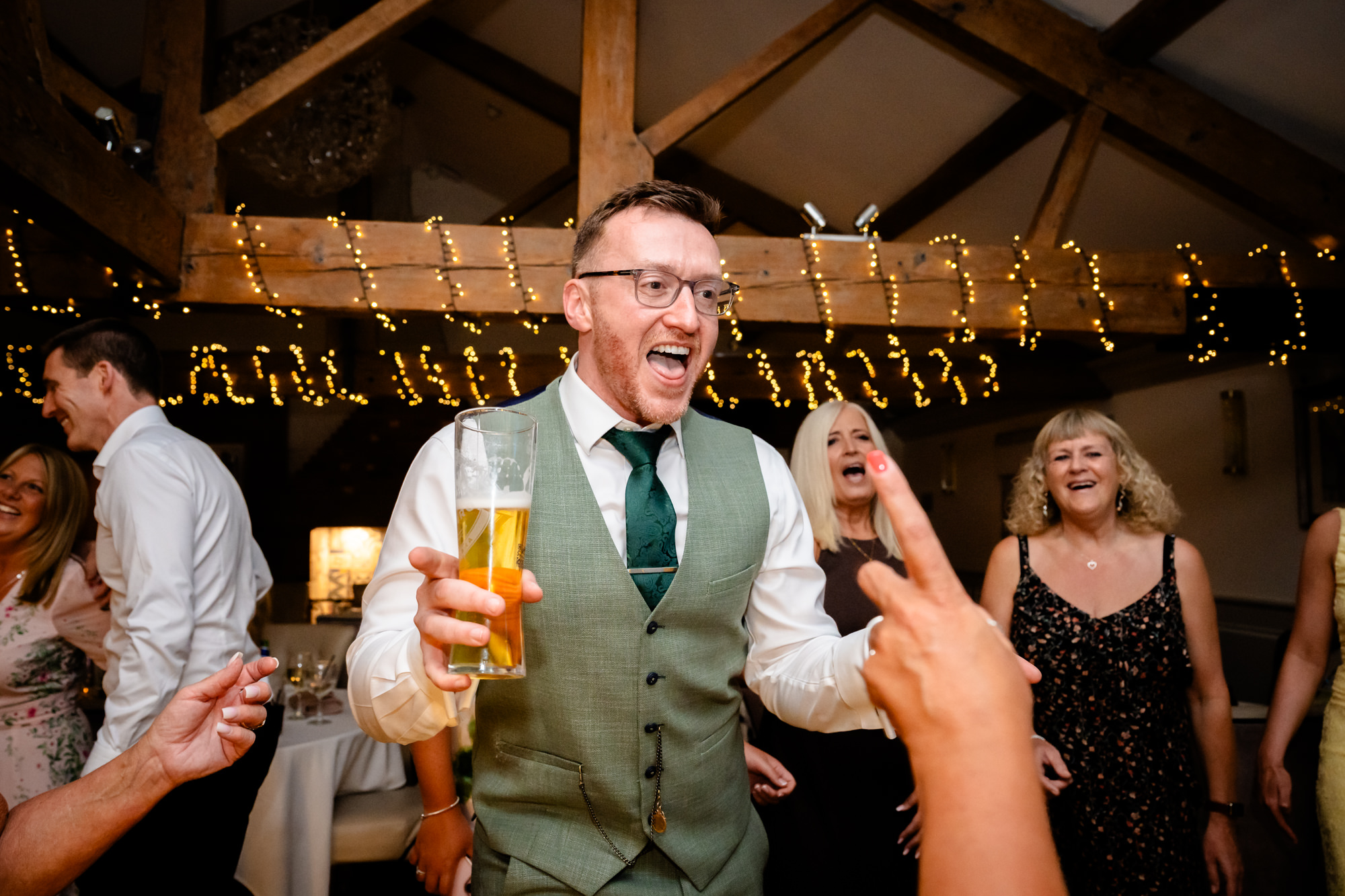 A groom in a green suit smiles broadly while holding a glass of beer, surrounded by friends who are dancing.