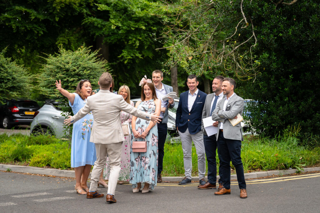 A group of six men and women, including the groom in the beige suit, stand in a line to welcome the groom