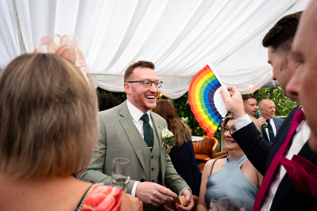 The groom in a green suit and green tie smiles widely as a guest to his right holds up a rainbow fan.
