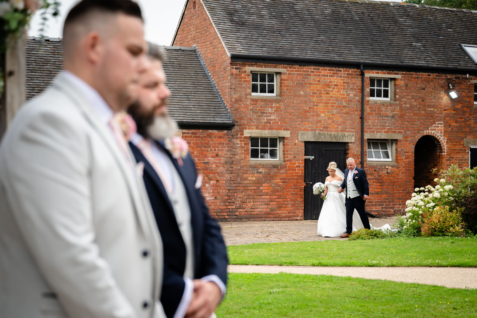 A long shot of the bride being escorted by her father across the lawn towards the ceremony area at Hanbury Barns.