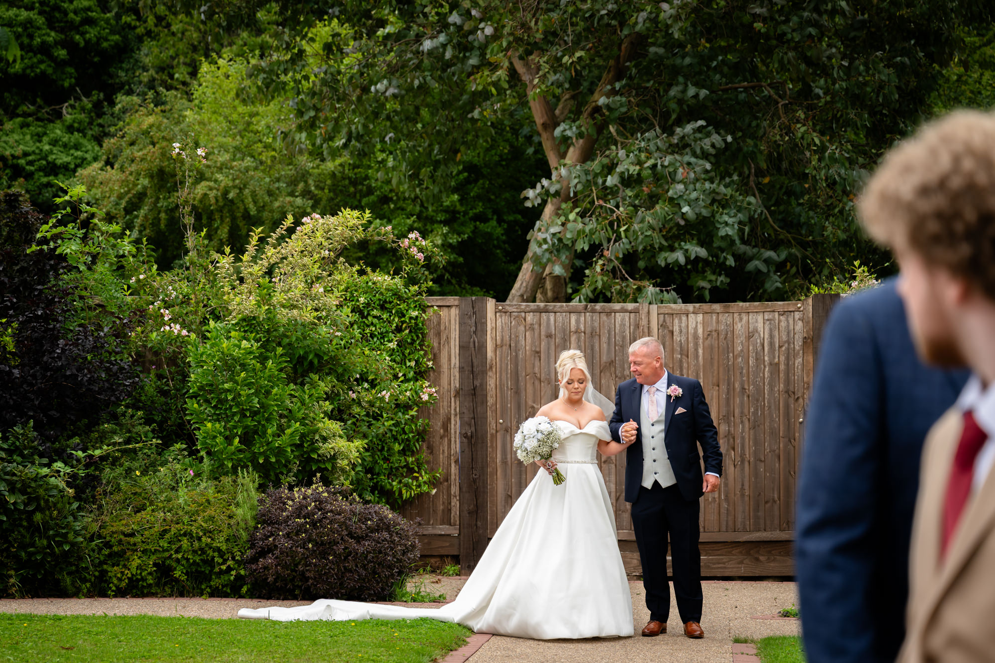 A wedding at Hanbury Barns perspective of the bride walking down the aisle with her father, captured between guests on a bright sunny day.