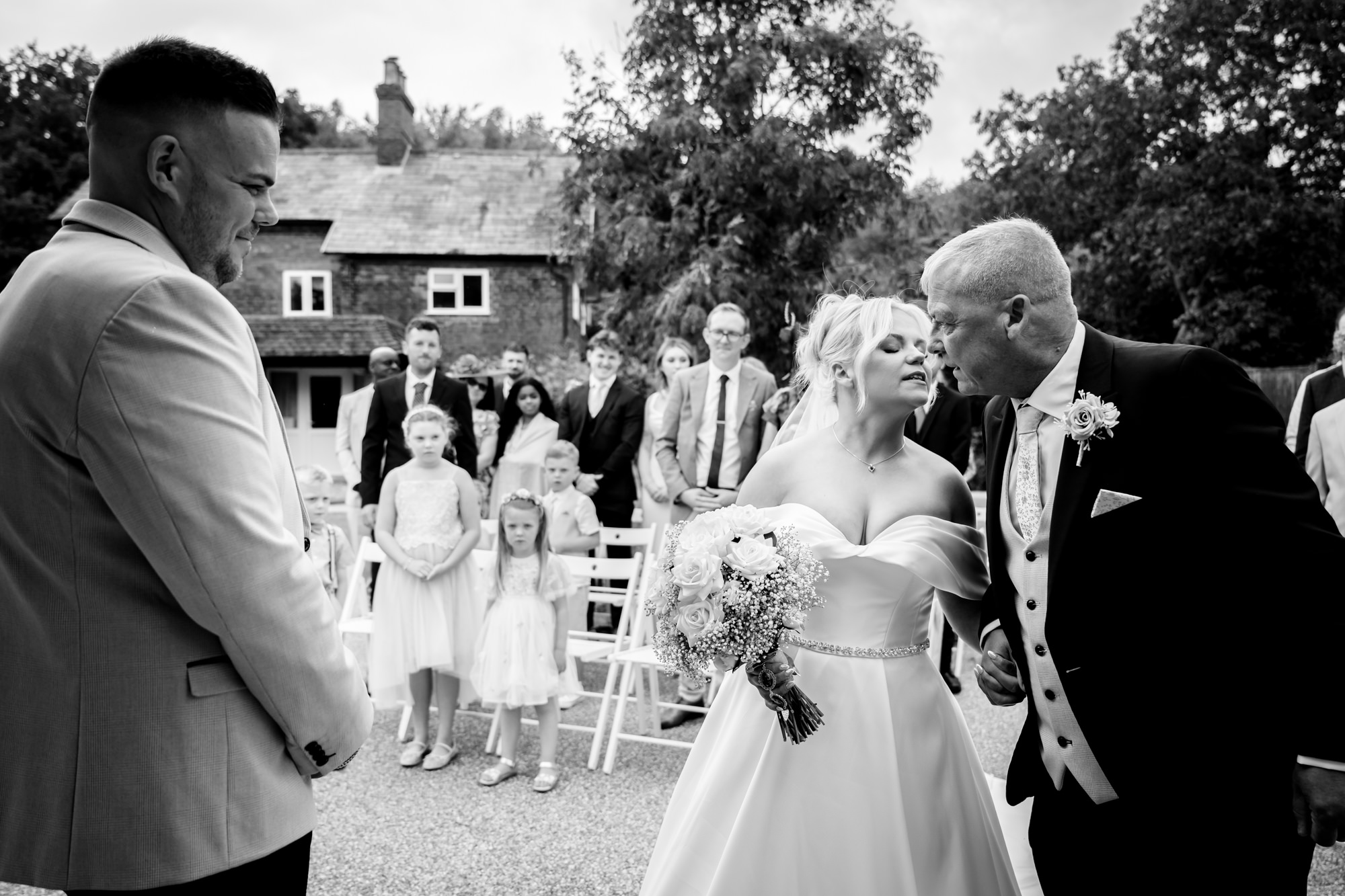 A black and white capture of the emotional exchange between the bride, her father, and the groom as she reaches the end of the aisle at Hanbury Barns.