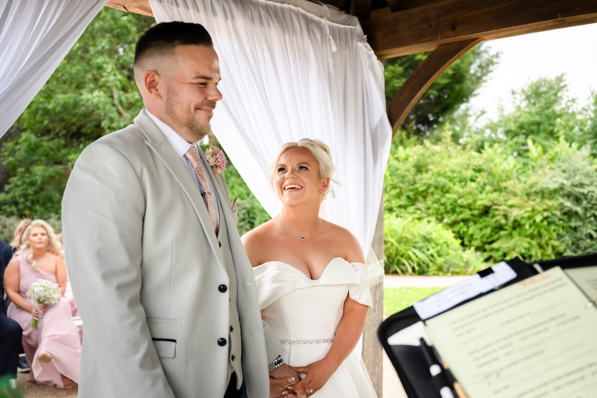 The wedding at Hanbury Barns bride and groom standing side-by-side in the gazebo, smiling and listening intently during the outdoor ceremony.
