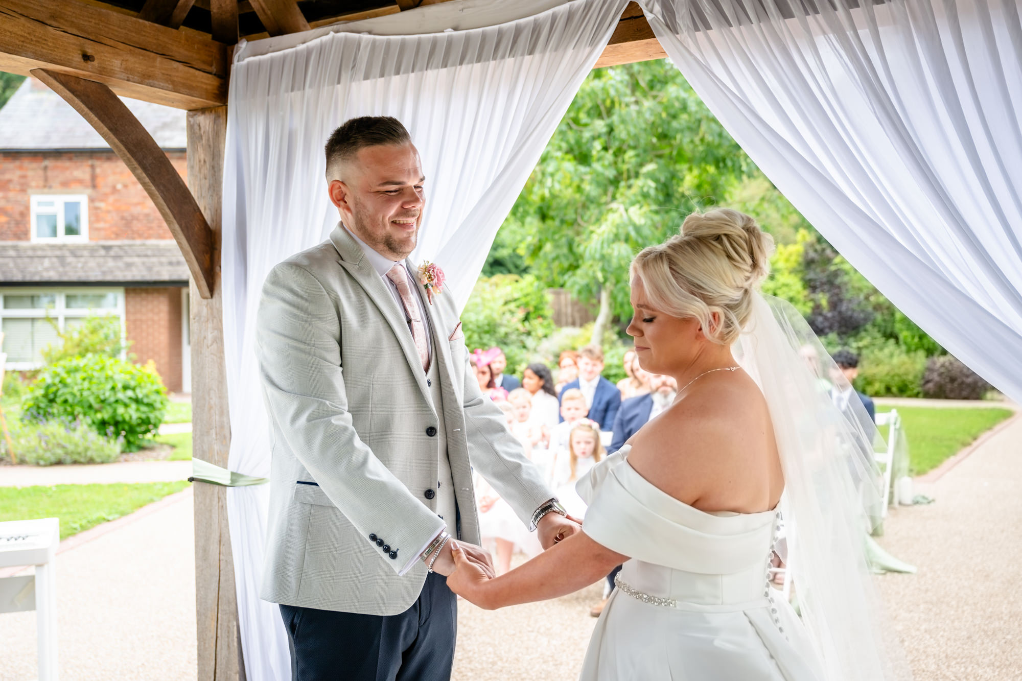 The couple holding hands under the white drapery of the outdoor structure, both looking happy during their vows at Hanbury Barns.