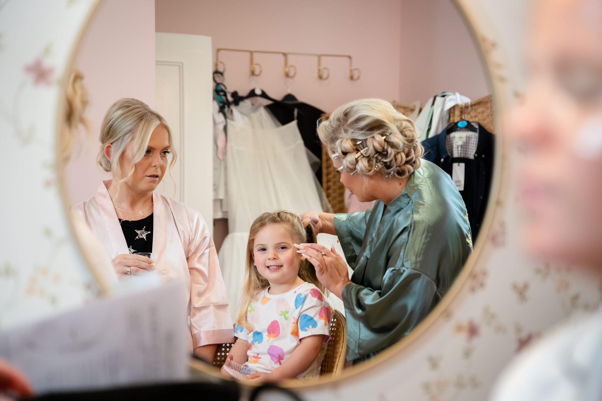 A flower girl smiling as a stylist works on her hair, reflected in a large round mirror inside Hanbury Barns.