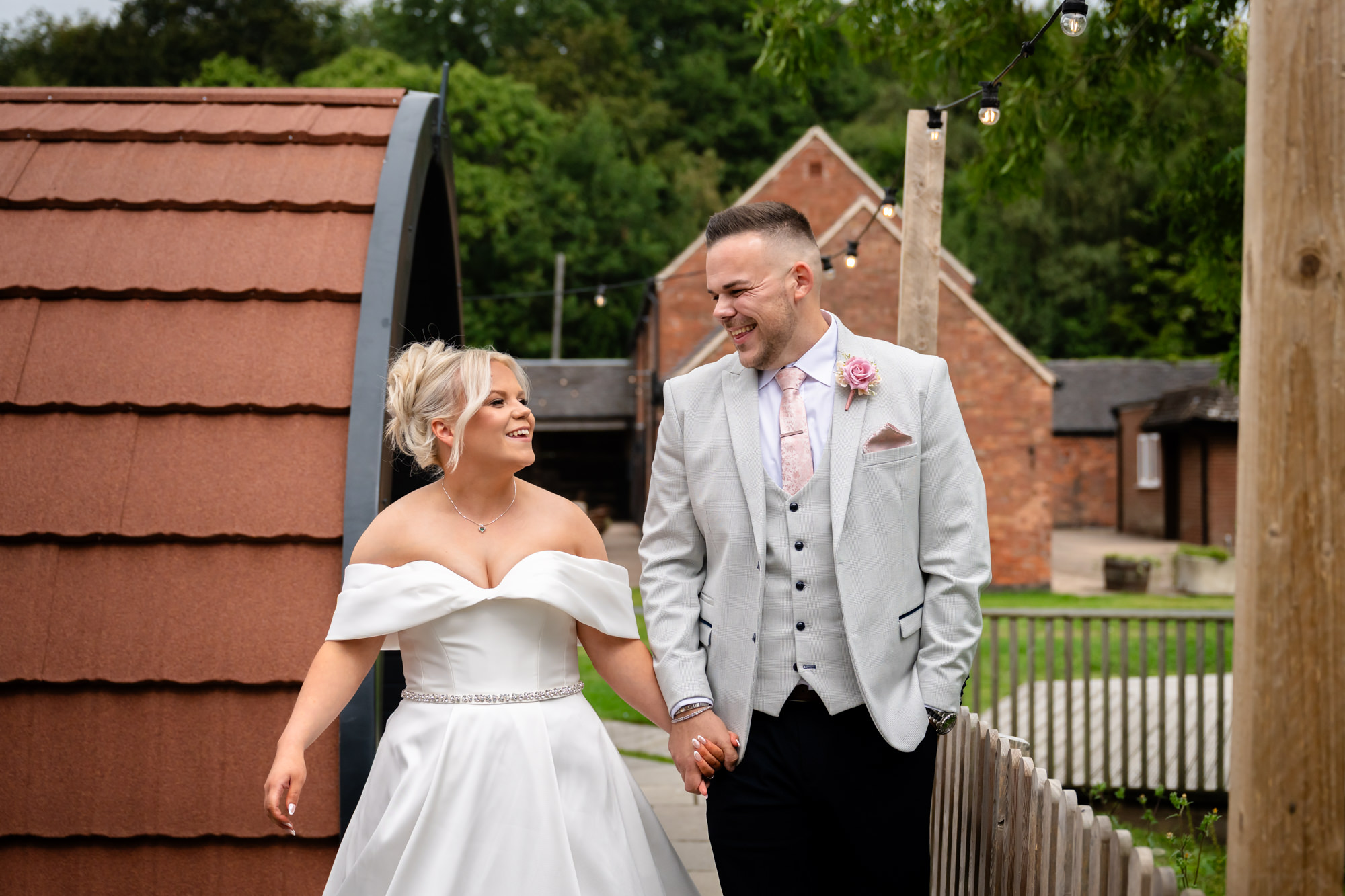 The bride and groom walking hand-in-hand and smiling at each other in front of the rustic wooden buildings at Hanbury Barns.